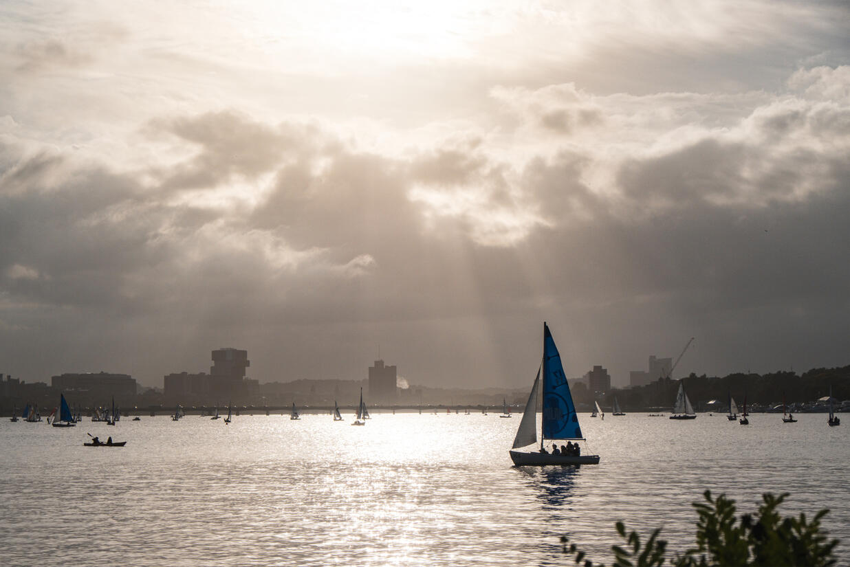 A sailboat glides on the calm Charles River. Many other boats and vessels litter the background. The Longfellow Bridge stretches across the city skyline, casting an orange glow on the water.