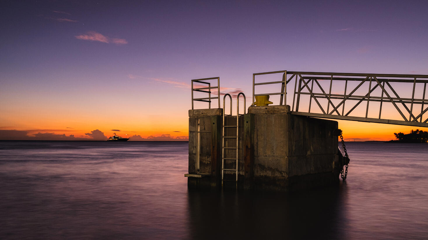 A wooden dock extending out into a calm body of water. The dock has a metal railing on either side, and a ladder attached to the side closest to the viewer. The ladder leads down into the glass like water. In the distance, there is a boat floating by again